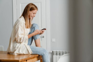 Evde masada otururken gülümseyen ve cep telefonu kullanan kızıl mutlu bir kız resmi