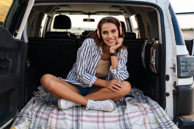 Genç, gülümseyen, kulaklıklı bir kadın arabanın arkasında oturuyor.