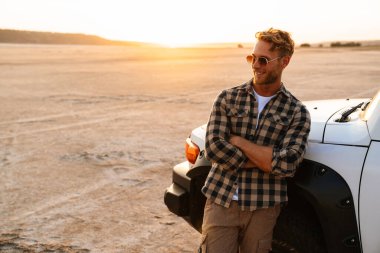 Yakışıklı, kendine güveni tam genç bir adam plajda arabasının kapısında dikilirken poz veriyor.