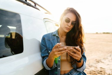 Plajda cep telefonu kullanarak arabaya yaslanan çekici genç bir kadın.