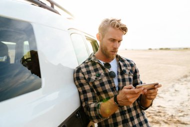 Plajda bir arabaya yaslanırken cep telefonu kullanan çekici bir genç.