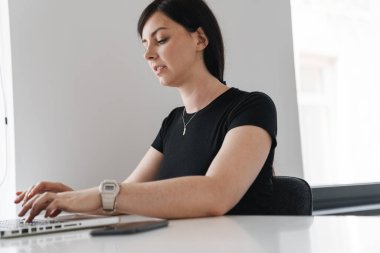 İşyerinde otururken dizüstü bilgisayarda çalışan konsantre genç bir kadın.