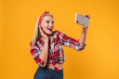 Sarı arka planda izole edilmiş cep telefonuyla gülümseyen ve selfie çeken mutlu poster kızı görüntüsü