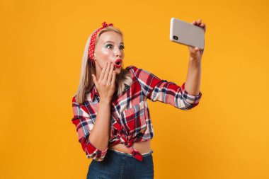 Sarı arka planda izole edilmiş cep telefonuyla gülümseyen ve selfie çeken şok olmuş bir kız resmi.