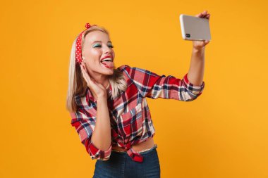 Dilini gösteren ve cep telefonundan selfie çeken mutlu bir poster kızı görüntüsü sarı arka planda izole edilmiş.