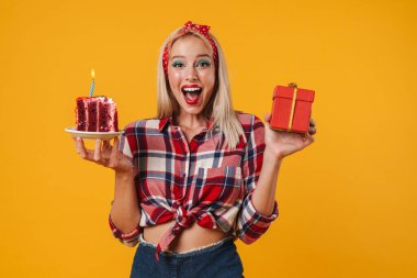 Sarı arka planda izole edilmiş hediye kutusu ve doğum günü pastasıyla poz veren heyecanlı çekici bir kız resmi