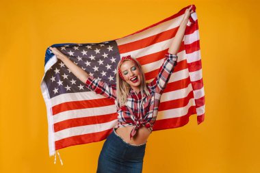 Amerikan bayrağıyla poz veren ve sarı arka planda izole bir şekilde gülümseyen güzel bir kız resmi.