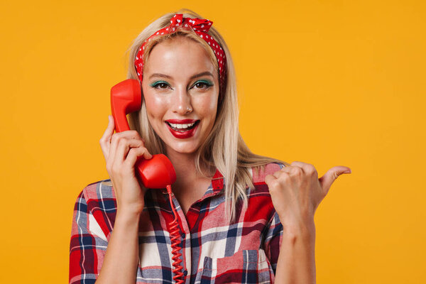 Изображение веселой девушки пинап указывая пальцем в сторону, позируя с телефоном изолированы на желтом фоне
