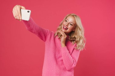 Pembe arka planda izole edilmiş cep telefonundan selfie çekerken gülümseyen mutlu sarışın kız görüntüsü