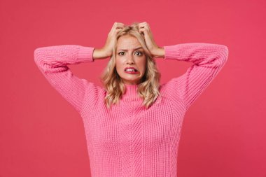 Kafası karışmış sarışın bir kız görüntüsü. Kameraya bakıyor ve pembe arka planda kafasını tutuyor.