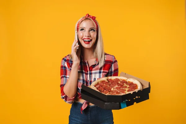 照片上 一个快乐迷人的女孩一边拿着披萨一边拿着手机在黄色的背景下聊天 — 图库照片