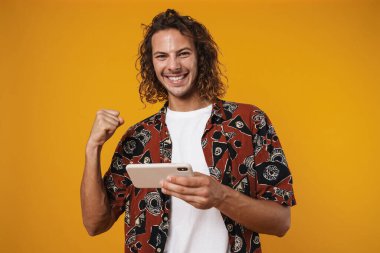 Sarı arka planda izole edilmiş cep telefonu kullanarak kazanan kıvırcık adamın fotoğrafı.