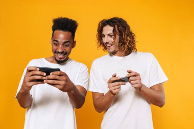 Gülümseyen çok kültürlü iki adam sarı arka planda izole edilmiş akıllı telefonlarda online oyunlar oynuyorlar.