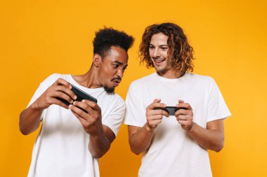 Odaklanmış çok kültürlü iki adam sarı arka planda izole edilmiş akıllı telefonlar üzerinde online oyunlar oynuyorlar.