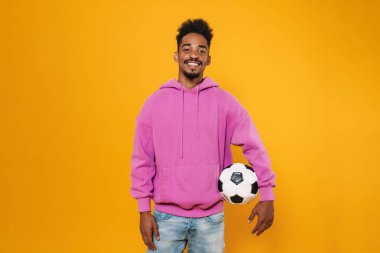 Sarı arka planda futbolla poz verirken gülümseyen mutlu Afrikalı Amerikalı adam.