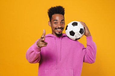 Mutlu Afro-Amerikalı adam sarı arka planda futbolla poz verirken kamerayı işaret ediyor.