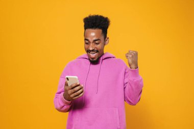 Heyecanlı Afrikalı Amerikalı adam kazanan hareketi yapıyor ve sarı arka planda izole edilmiş cep telefonu kullanıyor.