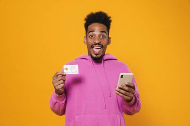 Sarı arka planda izole edilmiş kredi kartı ve cep telefonuyla poz veren Afro-Amerikan bir adam.
