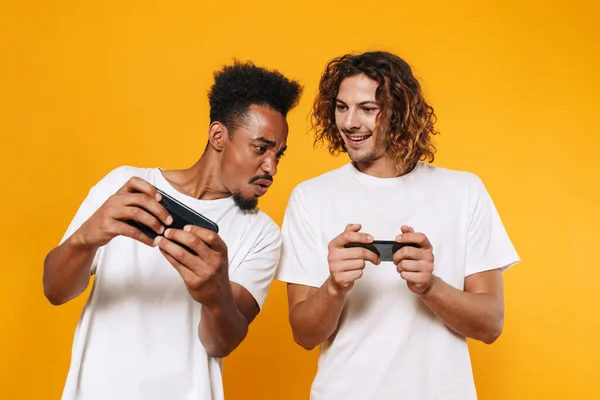 黄色の背景に隔離されたスマートフォンでオンラインゲームをプレイする多文化2人の男に焦点を当て — ストック写真