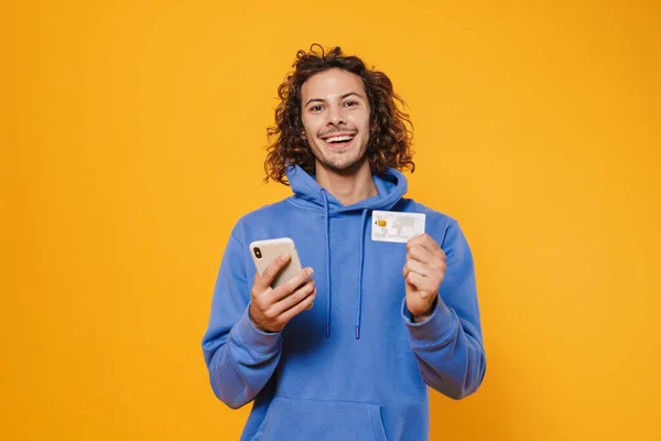 兴奋的高加索人在黄色背景下摆出手机和信用卡的姿势 — 图库照片