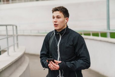 Stadyumda yürüyen, spor çantası taşıyan, cep telefonu kullanan kendinden emin genç bir adam.