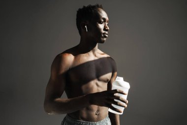 Gri arka planda izole edilmiş su içerken kulaklık kullanan Afro-Amerikan sporcusu.