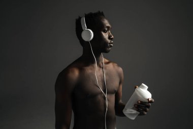 Gri arka planda izole edilmiş su içerken kulaklık kullanan üstsüz Afro-Amerikan sporcusu.