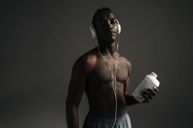 Gri arka planda izole edilmiş su içerken kulaklık kullanan üstsüz Afro-Amerikan sporcusu.
