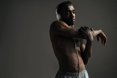 Gömleksiz Afro-Amerikan sporcusu, kulaklık takıp kollarını geriyor. Gri arka planda izole bir şekilde.