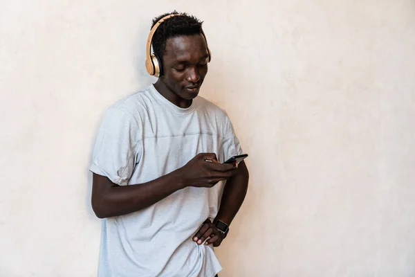 Молодой Африканский Американец Наушниками Держит Мобильник Стоя Над Стеной Помещении — стоковое фото