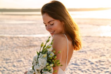 Güzel güneşli bir sabahta elinde buketle sahilde yürüyen neşeli, iyimser ve mutlu genç bir kadının fotoğrafı.