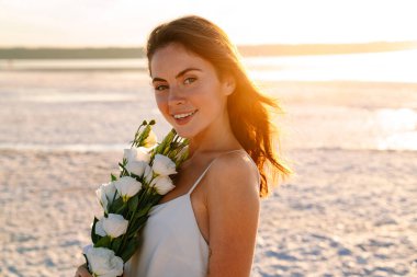 Güzel güneşli bir sabahta elinde buketle sahilde yürüyen neşeli, iyimser ve mutlu genç bir kadının fotoğrafı.