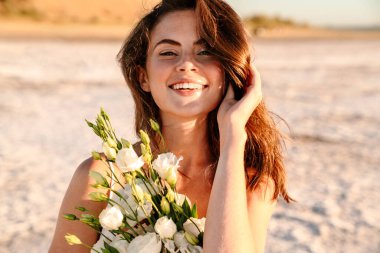 Güzel güneşli bir sabahta elinde buketle sahilde yürüyen neşeli, iyimser ve mutlu genç bir kadının fotoğrafı.