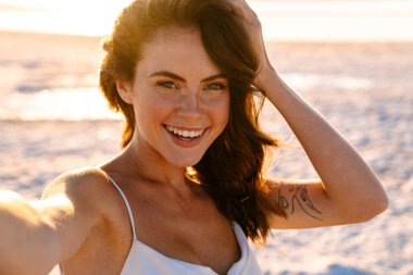 Gün batımında sahilde beklerken selfie çeken gülümseyen genç bir kadın.