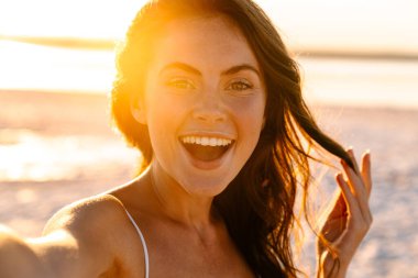 Gün batımında sahilde beklerken selfie çeken gülümseyen genç bir kadın.