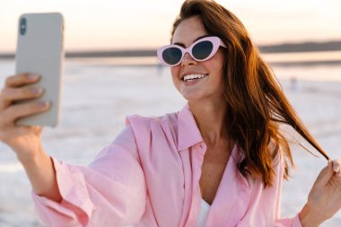 Gün batımında sahilde güneş gözlüğü takmış, selfie çeken neşeli genç bir kadın.