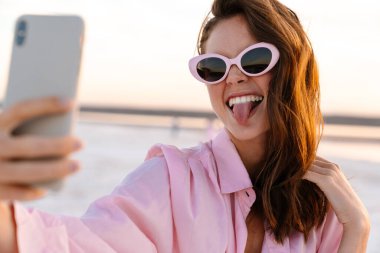 Gün batımında sahilde güneş gözlüğü takmış, selfie çeken neşeli genç bir kadın.