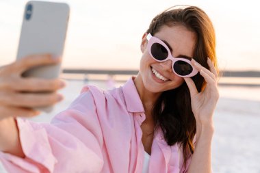 Gün batımında sahilde güneş gözlüğü takmış, selfie çeken neşeli genç bir kadın.