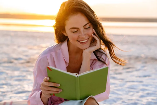 Sonriendo Hermosa Joven Leyendo Libro Mientras Está Sentada Playa — Foto de Stock