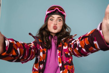 Snowboard kıyafeti giymiş neşeli genç bir kadın mavi sırt çantasından izole edilmiş bir selfie çekiyor.