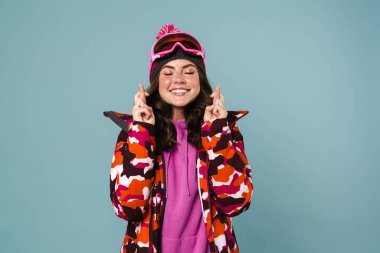 Snowboard ceketi giyen neşeli genç bir kadın şans dilemek için mavi arka planda beklemekte.