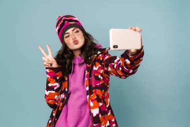 Snowboard kıyafeti giymiş neşeli genç bir kadın mavi sırt çantasından izole edilmiş bir selfie çekiyor.