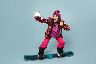 Neşeli sporcu kadın cep telefonuyla selfie çekerken snowboard 'da poz verirken mavi duvarda izole edilmiş.
