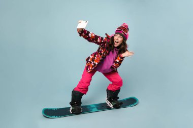 Neşeli sporcu kadın cep telefonuyla selfie çekerken snowboard 'da poz verirken mavi duvarda izole edilmiş.