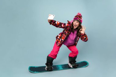 Neşeli sporcu kadın cep telefonuyla selfie çekerken snowboard 'da poz verirken mavi duvarda izole edilmiş.