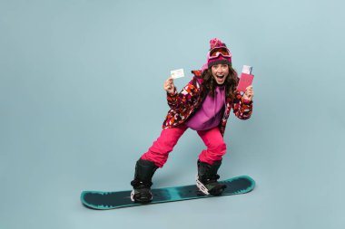 Heyecanlı sporcu kadın, pasaportu ile snowboard 'da poz veriyor. Mavi duvarda izole edilmiş, kredi kartı gösteriyor.