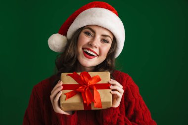 Noel Baba şapkalı sevimli kız Noel hediyesiyle poz veriyor yeşil arka planda.