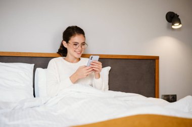 Güzel mutlu kız evde kötü otururken cep telefonu kullanıyor ve gülümsüyor.