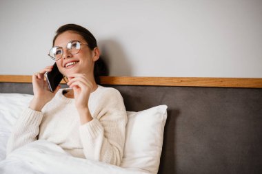 Güzel, neşeli bir kız cep telefonuyla konuşuyor ve evde kötü otururken gülümsüyor.