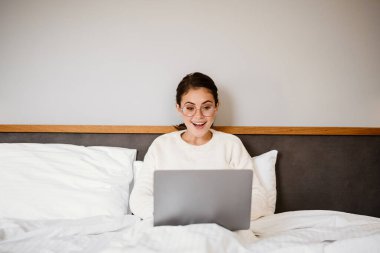 Güzel, neşeli kız evde kötü otururken gülümsüyor ve laptop kullanıyor.